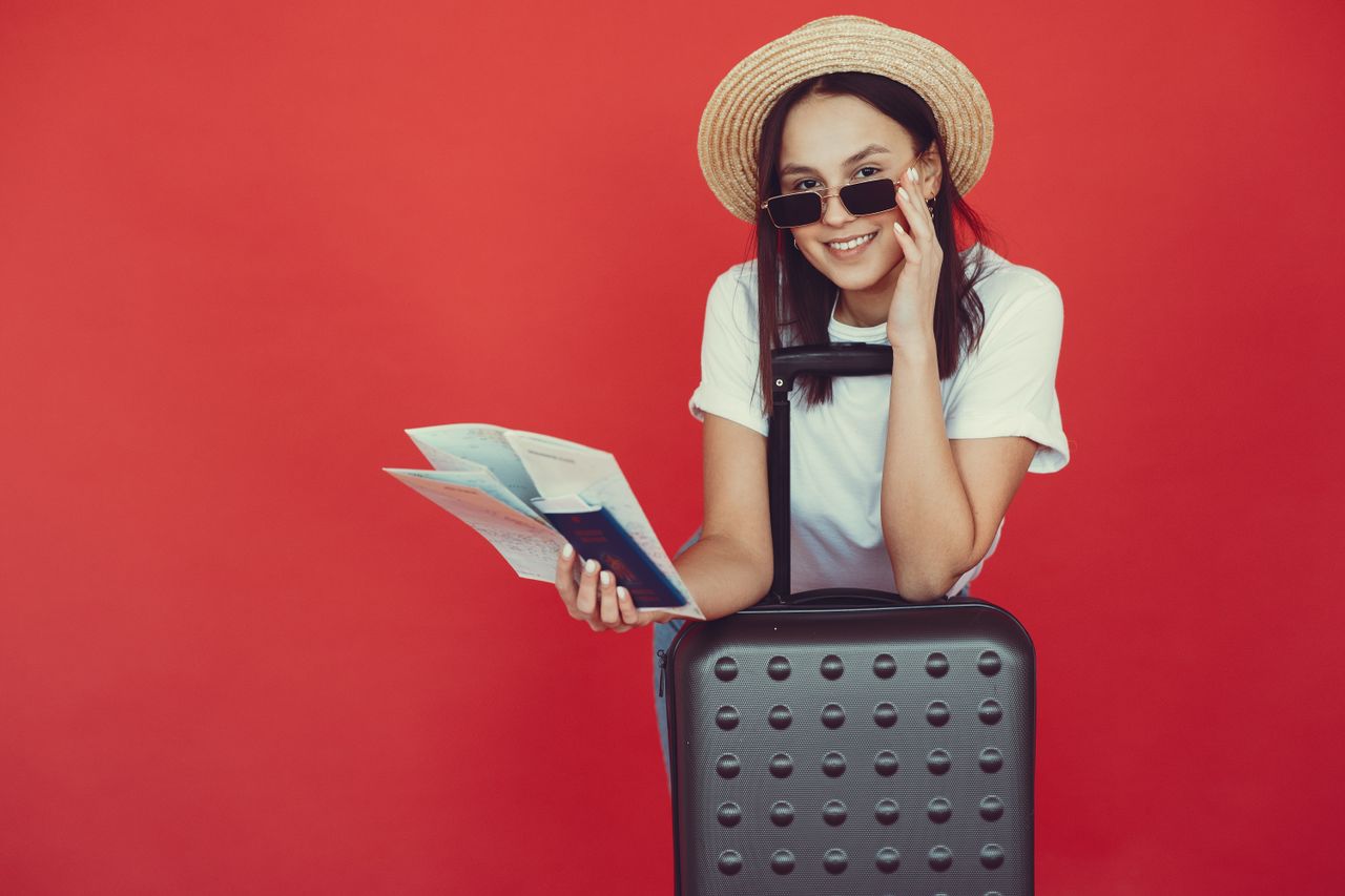services-02 Delighted young woman in straw hat in casual clothes leaning on suitcase and checking tickets before traveling on red background