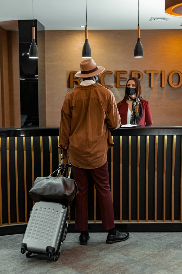 why-choose-us Man with luggage checking in at hotel reception. Staff wearing mask, indoors, travel theme.
